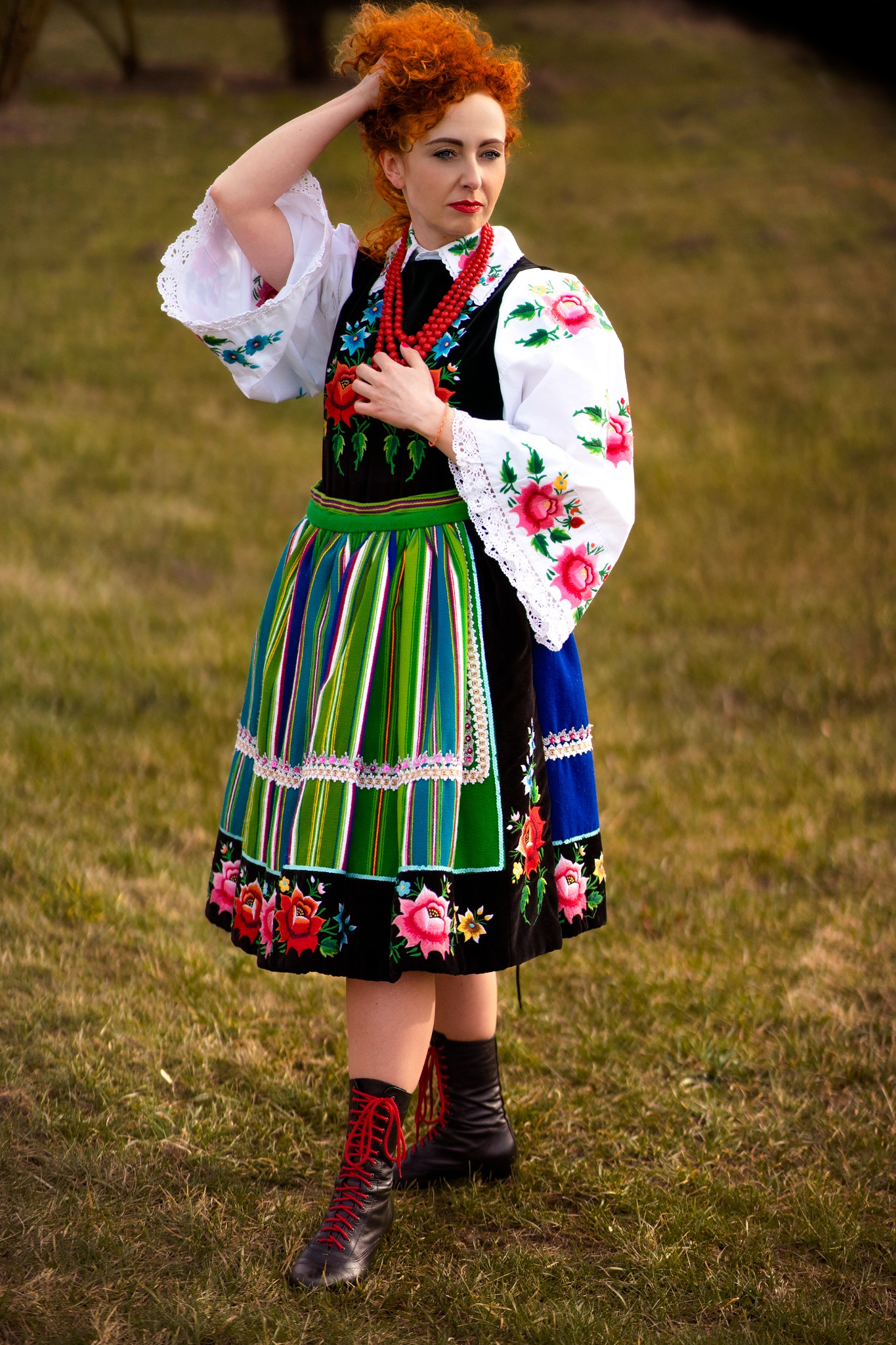 Kobieta w tradycyjnym stroju ludowym z czerwonymi koralami i butami sznurowanymi czerwoną wstążką, pozująca na łące.
