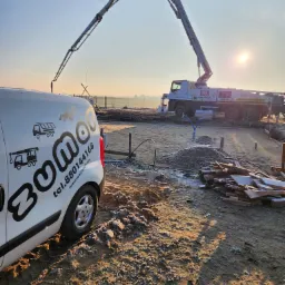 Wczesnoporanny widok na plac budowy w Postomino z pompą do betonu w tle, widoczny wykop pod fundamenty i auto firmy z logo.