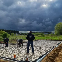Trzech robotników wiąże stal zbrojeniową na przygotowanym fundamencie płyty, widoczne szalunki, w tle burzowe chmury nad polami.