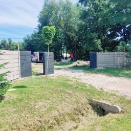 pphu. JN. Grek-Bud Grzegorz Gąsiorek - Nowoczesny, modułowy płot betonowy w odcieniach szarości, z otwartą bramą wjazdową. Widoczny fragment posesji z zielenią i zaparkowany samochód dostawczy.