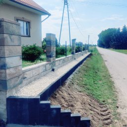 pphu. JN. Grek-Bud Grzegorz Gąsiorek - Nowoczesny murek oporowy z elementami betonowymi i kamieniami ozdobnymi, oddzielający posesję od drogi. Widoczne słupki i podmurówka w trakcie budowy.