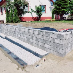 pphu. JN. Grek-Bud Grzegorz Gąsiorek - Nowoczesny murek oporowy z szarej kostki, schody z białym grysem, widoczne narzędzia i materiały budowlane. Prace w toku, dom w tle.