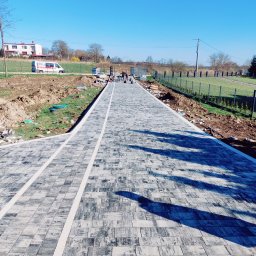 pphu. JN. Grek-Bud Grzegorz Gąsiorek - Długa, szara ścieżka z kostki brukowej z białymi pasami, prowadząca w kierunku ogrodzenia i domu w oddali, w słoneczny dzień. Widoczne prace wykończeniowe.