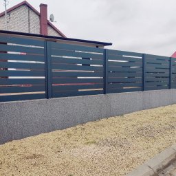 Nowoczesne, poziome przęsła ogrodzeniowe w kolorze grafitowym, zamontowane na betonowej podmurówce. Tło stanowi budynek mieszkalny i wysypany kamień ozdobny.