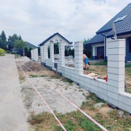 pphu. JN. Grek-Bud Grzegorz Gąsiorek - Wznoszenie ogrodzenia z prefabrykatów betonowych na posesji. Widoczne słupki i fragmenty muru, teren zabezpieczony taśmą. W tle domy jednorodzinne.