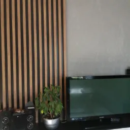 Drewniane lamele pionowe na czarnym tle, stanowiące dekoracyjny element ściany w salonie, obok szarego tynku dekoracyjnego. Poniżej telewizor Samsung, głośniki LG oraz roślina doniczkowa...