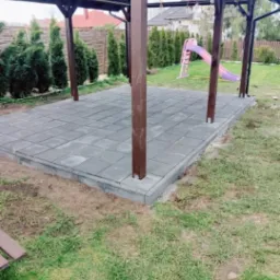 Szara kostka brukowa ułożona pod drewnianą altaną w ogrodzie, widoczna zieleń trawy i zjeżdżalnia w tle.