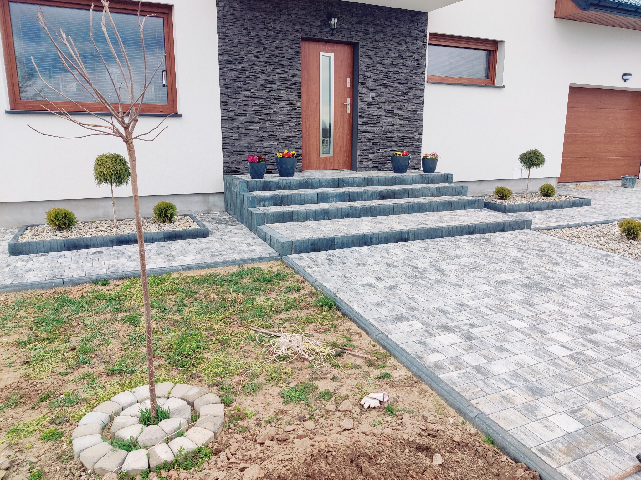 Ułożona kostka brukowa tworząca ścieżkę i schody przed nowoczesnym domem z elementami dekoracyjnymi, takimi jak kwiaty w donicach i małe drzewka.