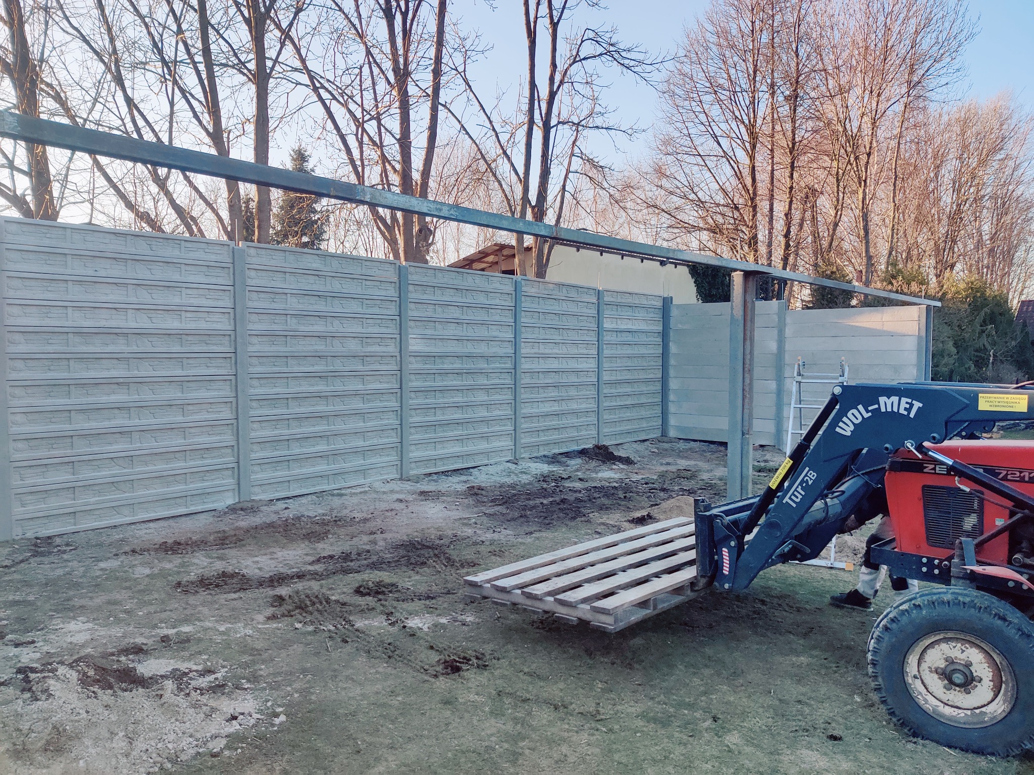 Montaż prefabrykowanego ogrodzenia betonowego z użyciem traktora z ładowaczem czołowym. Widoczne panele ogrodzeniowe i metalowa konstrukcja nośna.