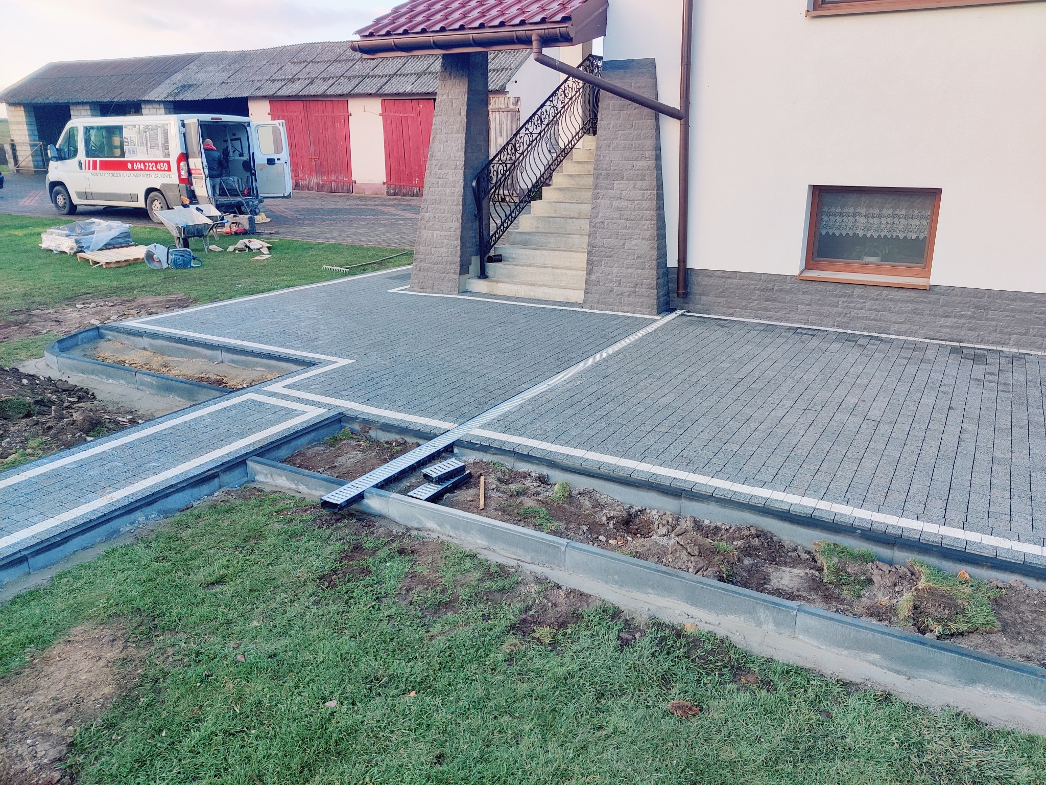 Ułożona kostka brukowa w szarym odcieniu, z białymi obrzeżami, tworząca ścieżkę i plac przed domem. Widoczne odwodnienie liniowe i częściowo wykopana ziemia pod trawnik.