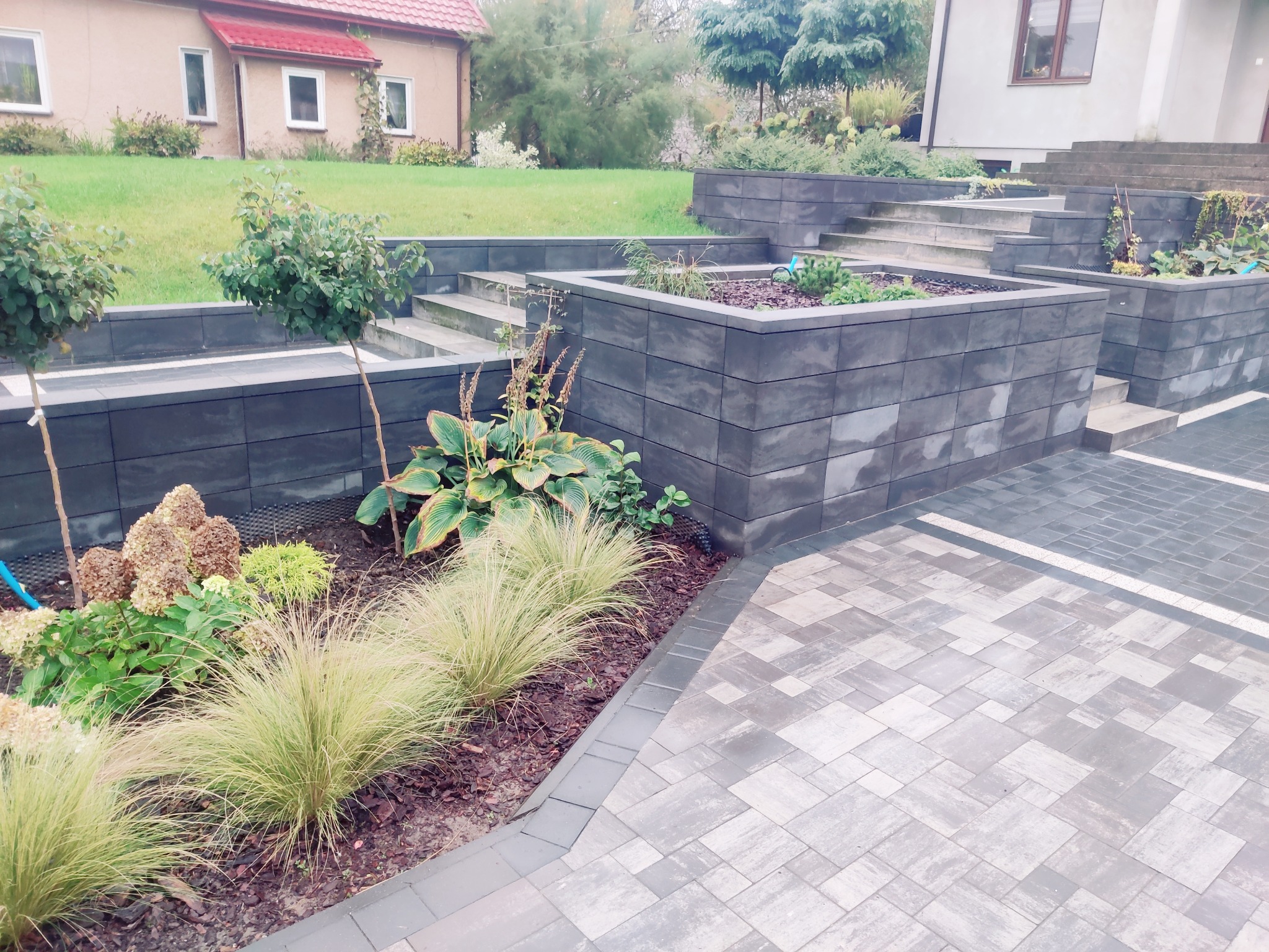 Nowoczesna aranżacja ogrodu z kostki brukowej i podwyższonych rabat z szarej kostki. Schody prowadzą do domu. Elementy zieleni i roślinności.