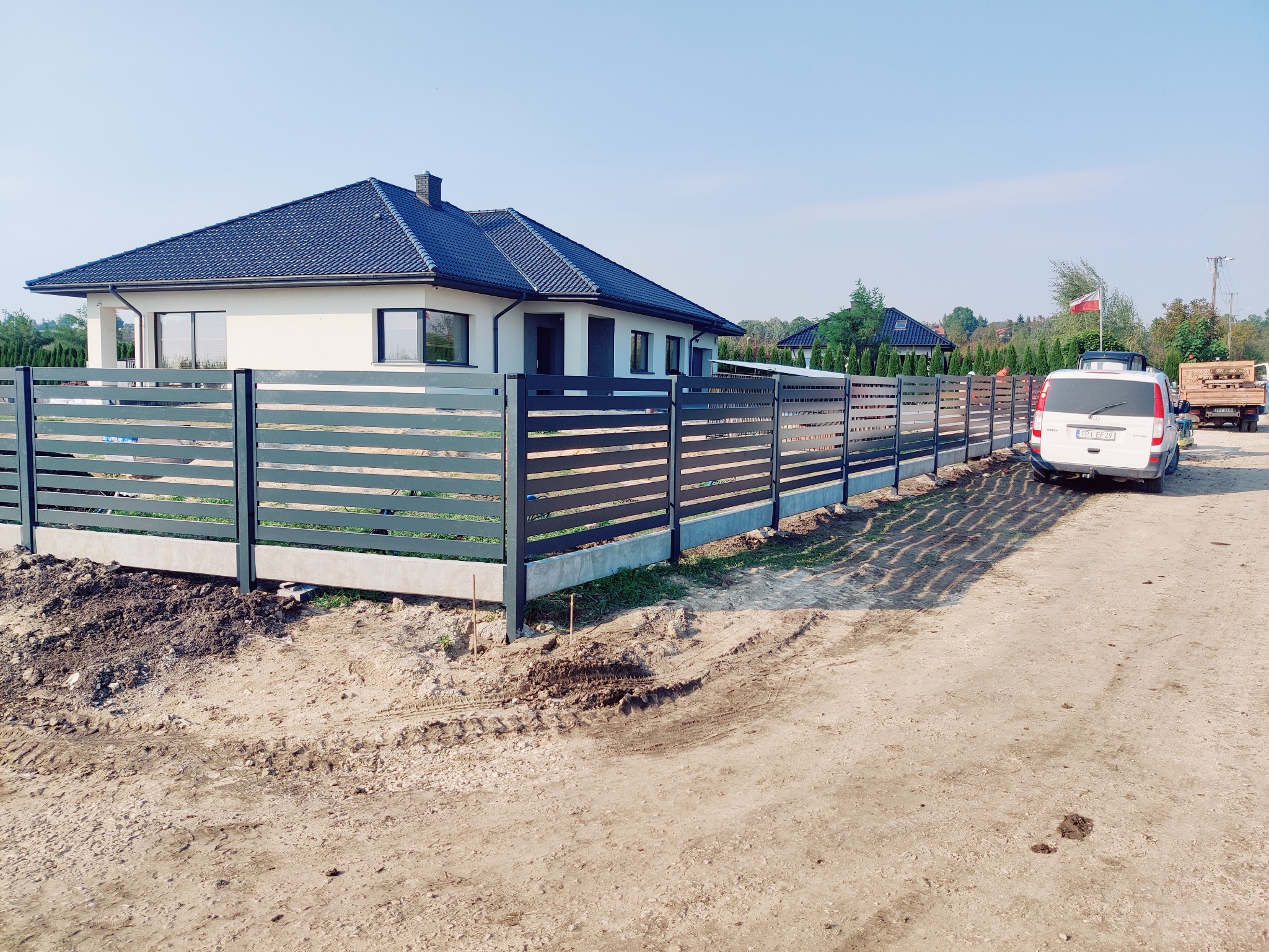 Nowoczesne, poziome ogrodzenie panelowe w kolorze grafitowym, z betonową podmurówką, otaczające posesję z domem jednorodzinnym. Widoczny samochód dostawczy i teren po pracach ziemnych.
