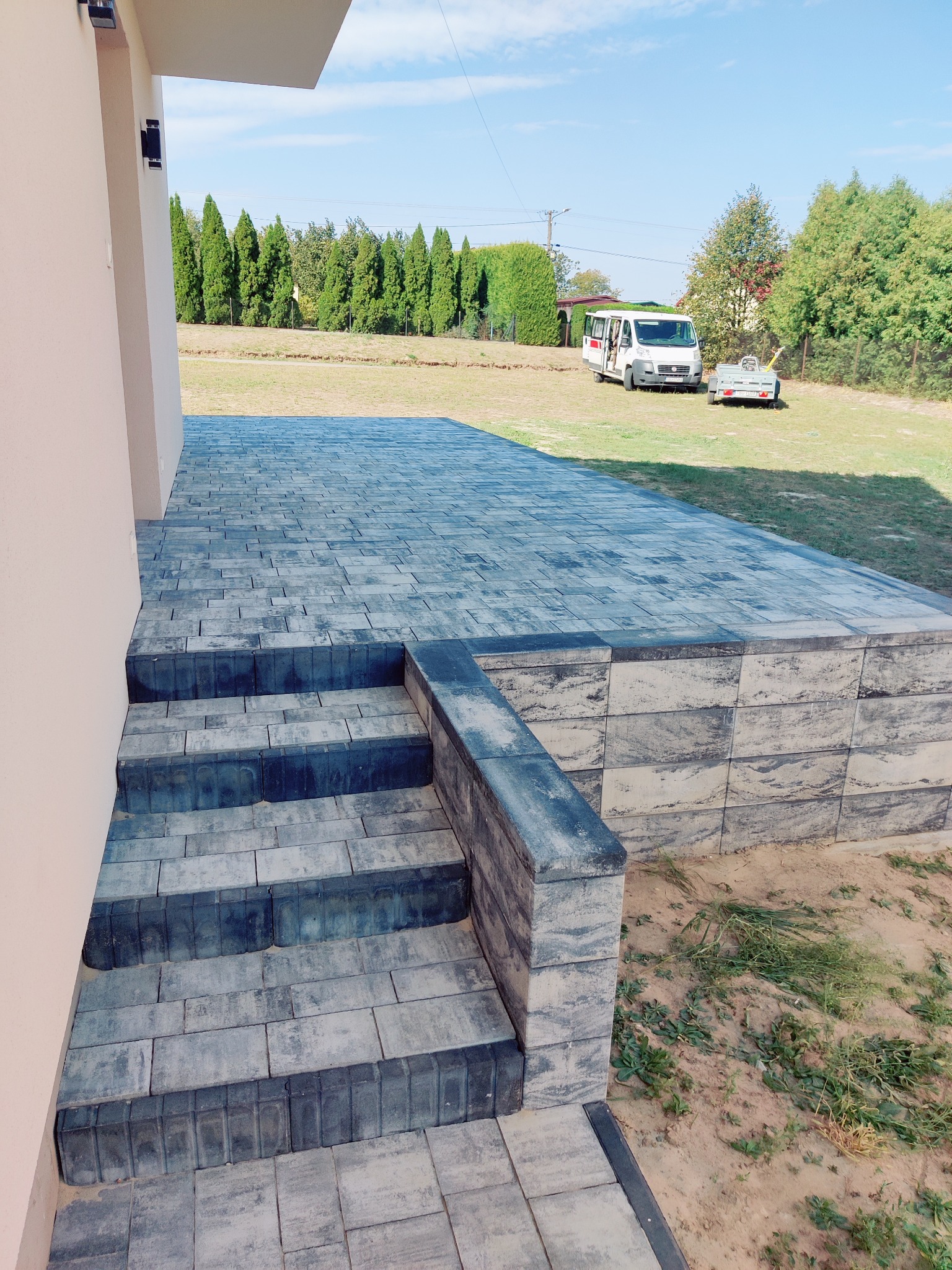 Ułożona kostka brukowa w odcieniach szarości tworząca schody i podest przy budynku. W tle trawnik, drzewa i zaparkowany bus z przyczepką. Słoneczny dzień.