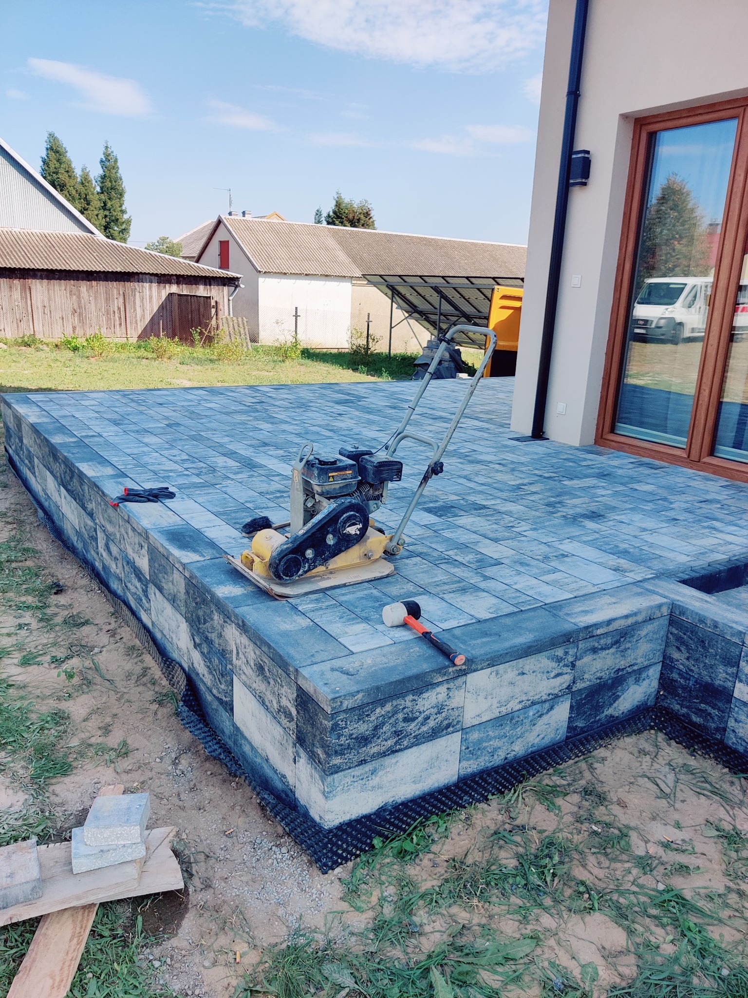 Niebiesko-szara kostka brukowa ułożona na tarasie przy domu. Na powierzchni ubijarka, młotek i rękawice robocze. Widoczne warstwy podbudowy.