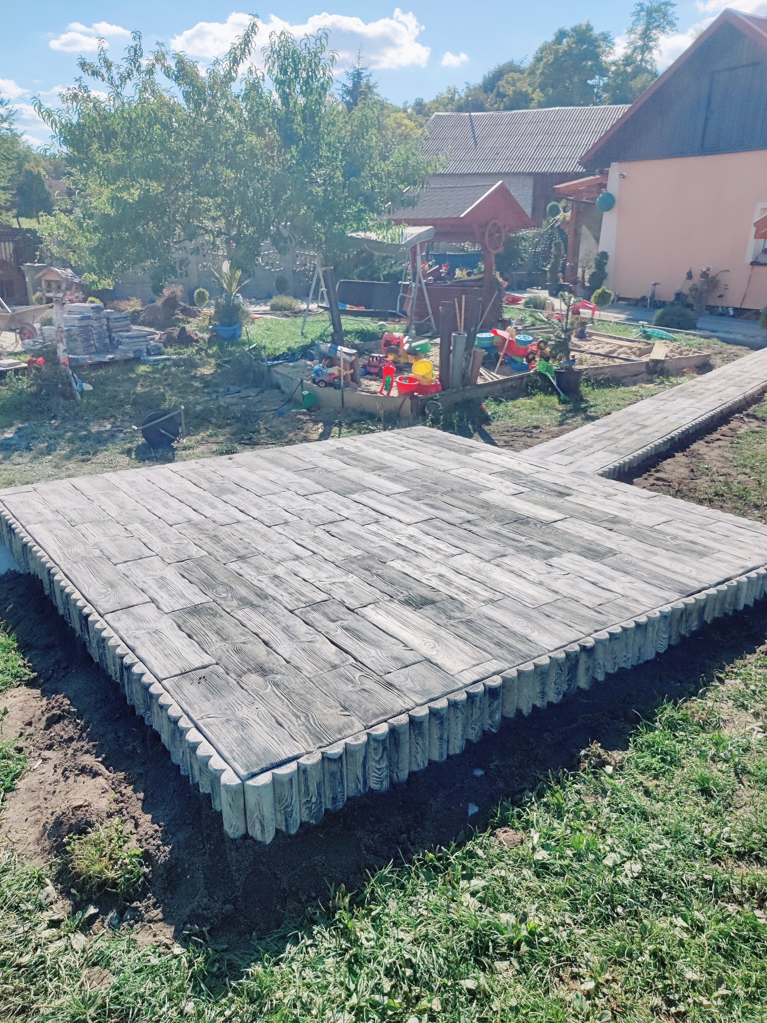 Ułożona kostka brukowa imitująca drewno, tworząca taras i ścieżkę w ogrodzie. Krawędzie wykończone elementami w kształcie drewnianych bali. Słoneczny dzień.