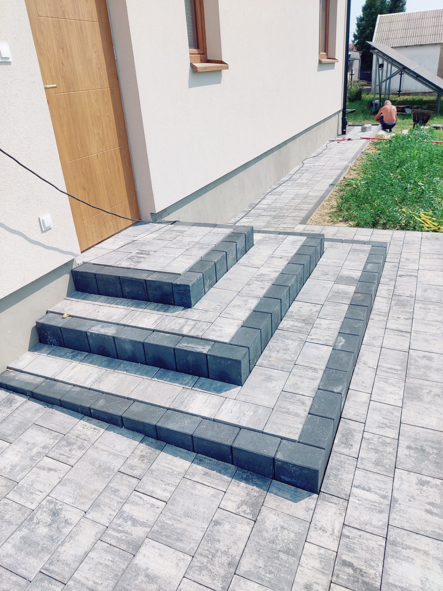 Ułożona kostka brukowa tworząca schody wejściowe do domu, z widocznym fragmentem chodnika i pracującym mężczyzną w tle. Szara kostka z ciemniejszym obramowaniem.