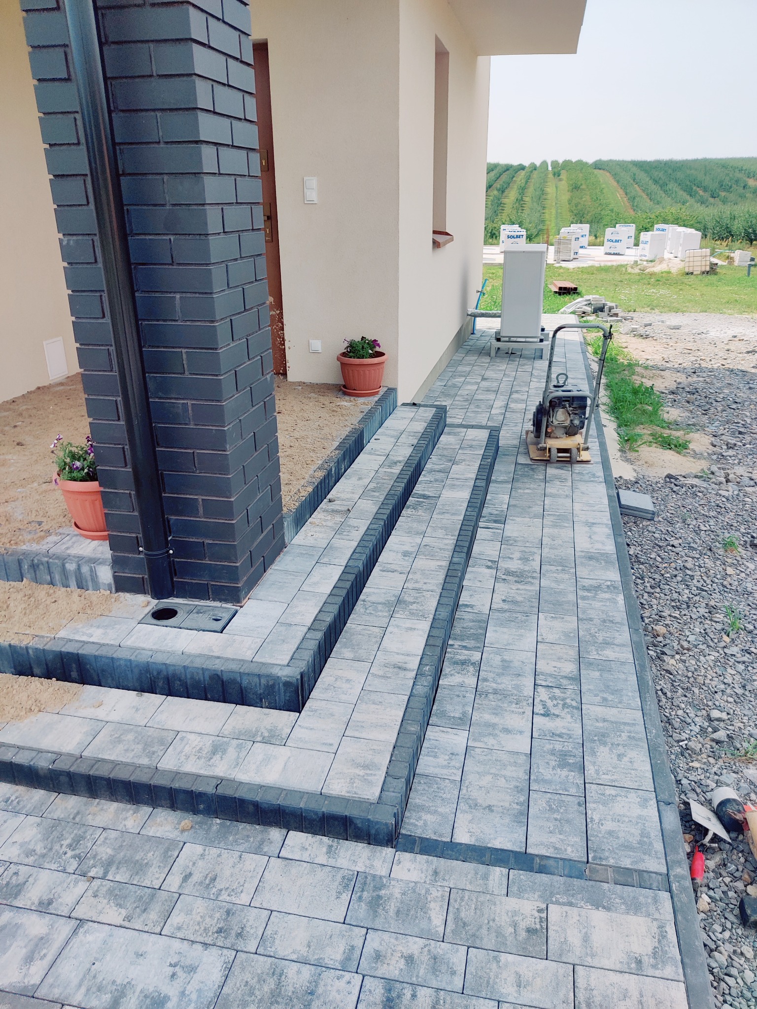 Ułożona kostka brukowa tworząca schody wejściowe do domu, z widoczną ubijarką i elementami wykończeniowymi. W tle budynek i zieleń.