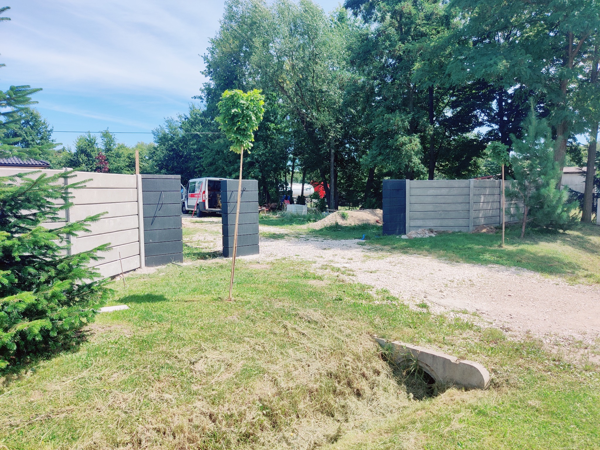 Nowoczesny, modułowy płot betonowy w odcieniach szarości, z otwartą bramą wjazdową. Widoczny fragment posesji z zielenią i zaparkowany samochód dostawczy.