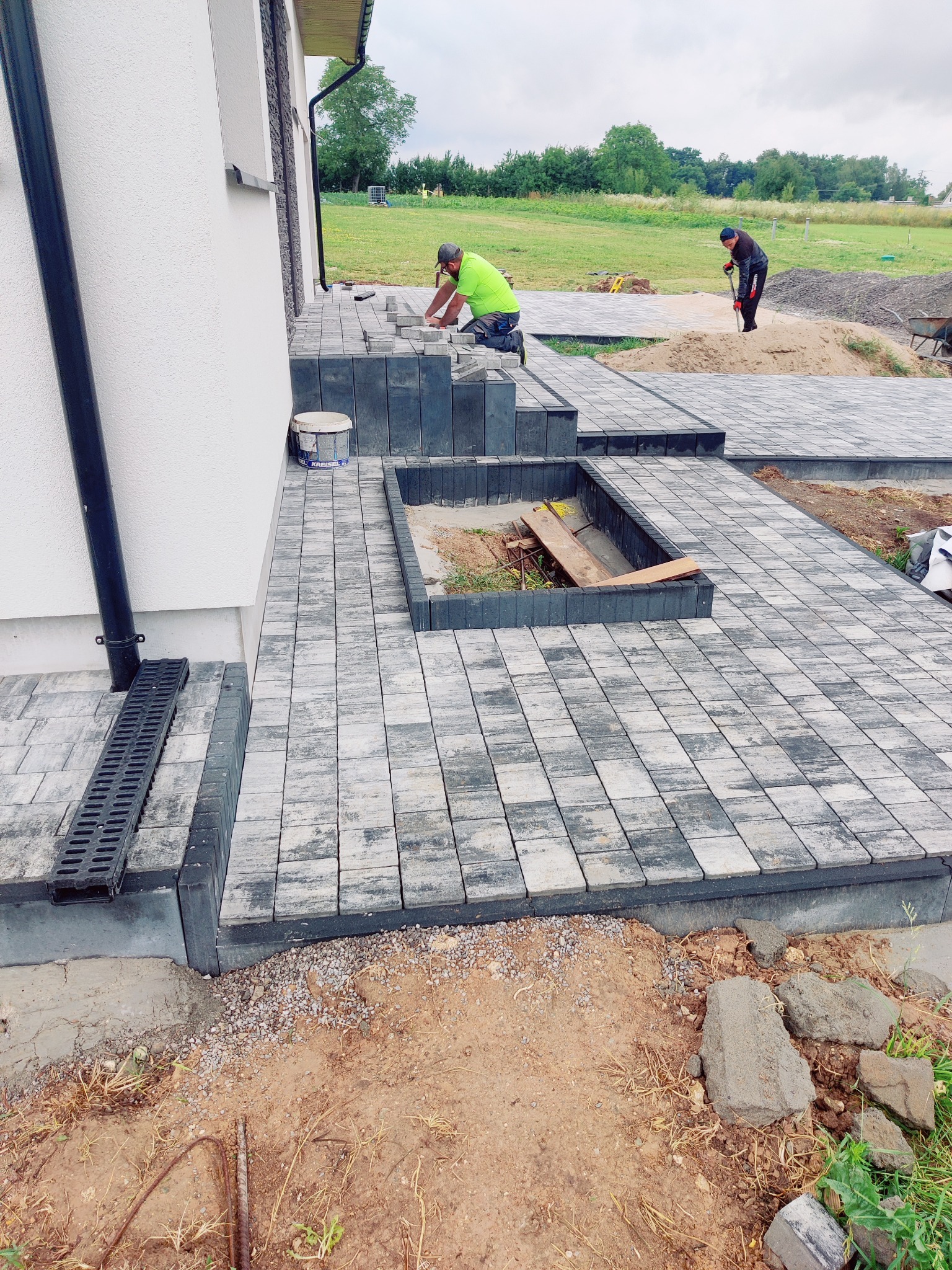 Układanie kostki brukowej na tarasie domu. Widoczni dwaj pracownicy podczas pracy. Kostka w odcieniach szarości, ułożona w regularny wzór. W tle zielona trawa i drzewa.