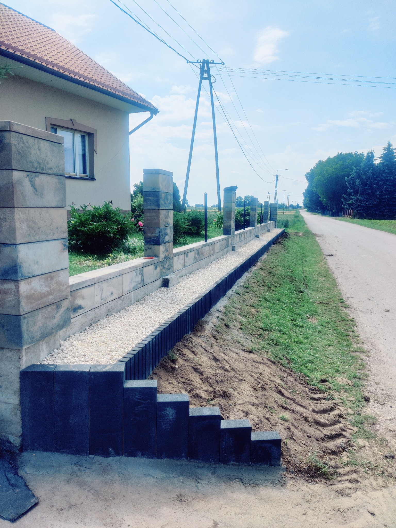Nowoczesny murek oporowy z elementami betonowymi i kamieniami ozdobnymi, oddzielający posesję od drogi. Widoczne słupki i podmurówka w trakcie budowy.