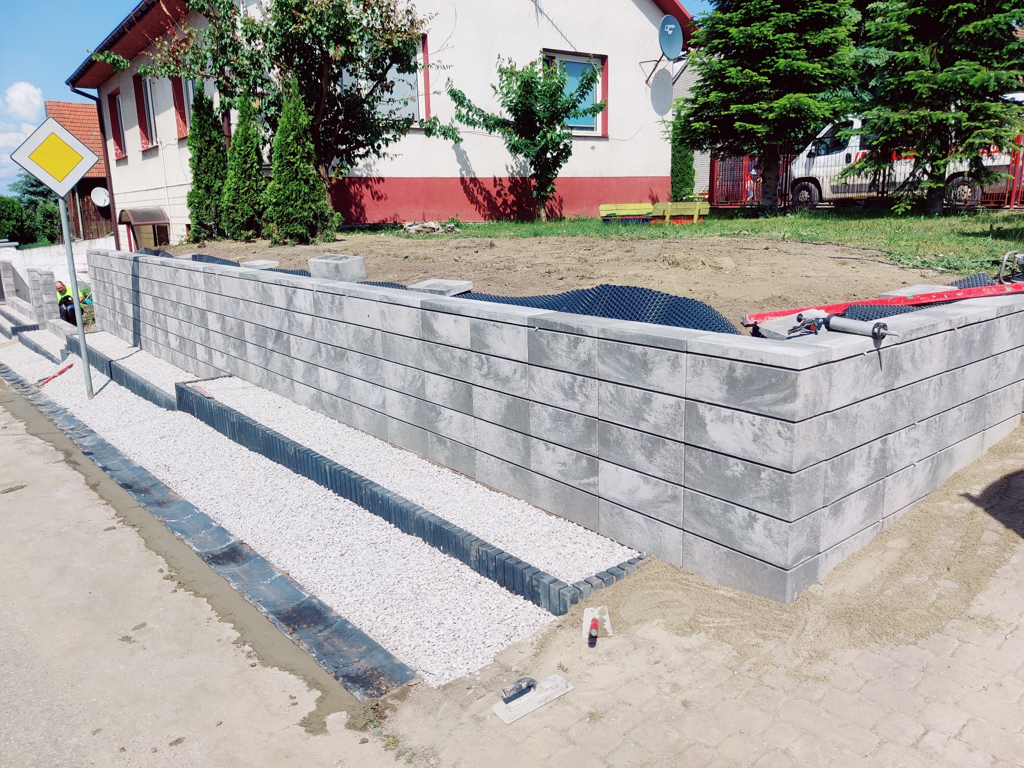Nowoczesny murek oporowy z szarej kostki, schody z białym grysem, widoczne narzędzia i materiały budowlane. Prace w toku, dom w tle.