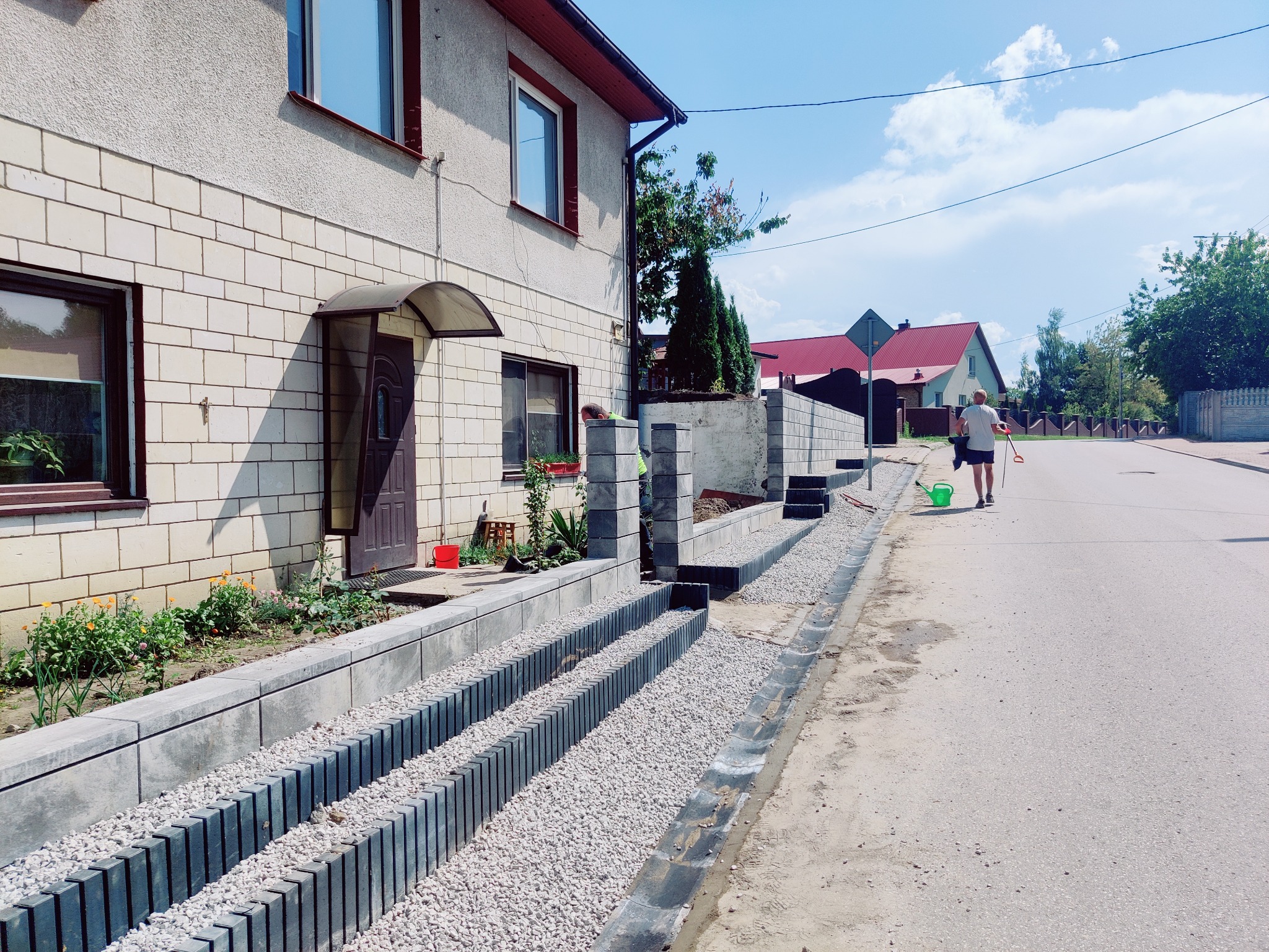Ukończona aranżacja terenu przed domem: kaskadowe rabaty z szarej kostki, wypełnione białym żwirem i zielenią. W tle budynek z jasnej cegły i osoba z konewką.