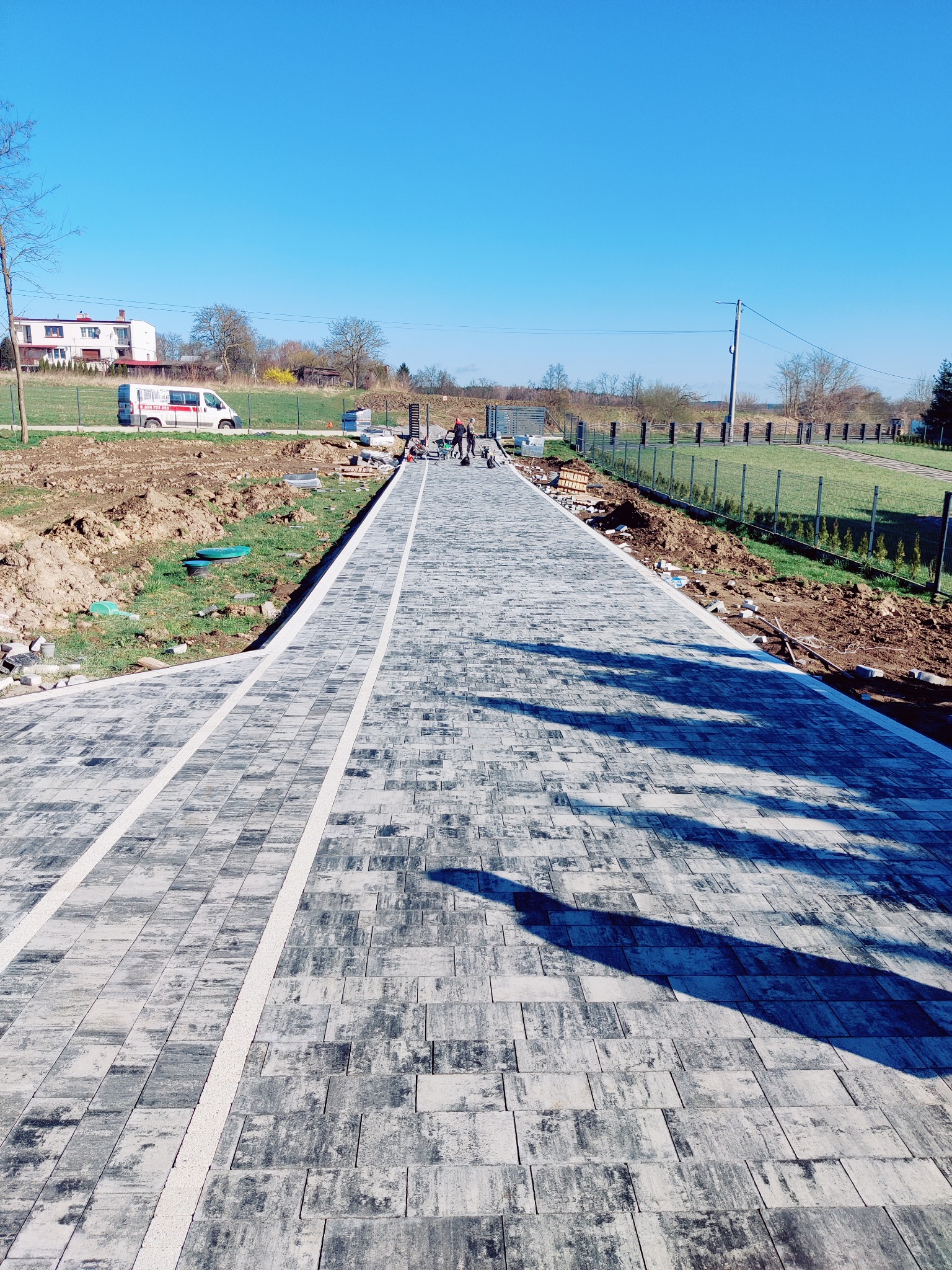 Długa, szara ścieżka z kostki brukowej z białymi pasami, prowadząca w kierunku ogrodzenia i domu w oddali, w słoneczny dzień. Widoczne prace wykończeniowe.