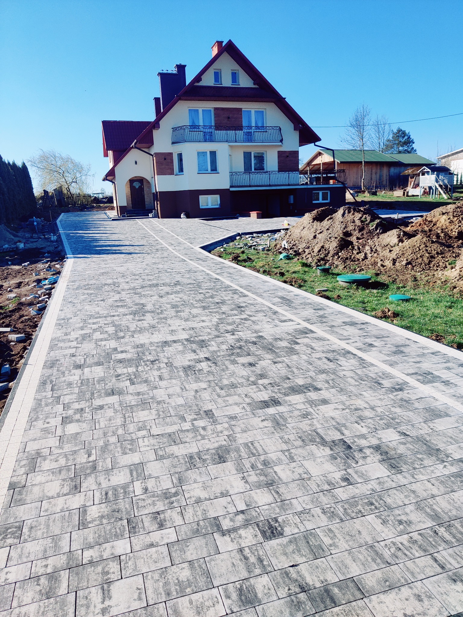 Ułożona kostka brukowa w odcieniach szarości tworzy ścieżkę prowadzącą do domu z balkonami. Widoczne przygotowania do dalszych prac ziemnych i fragment ogrodu.