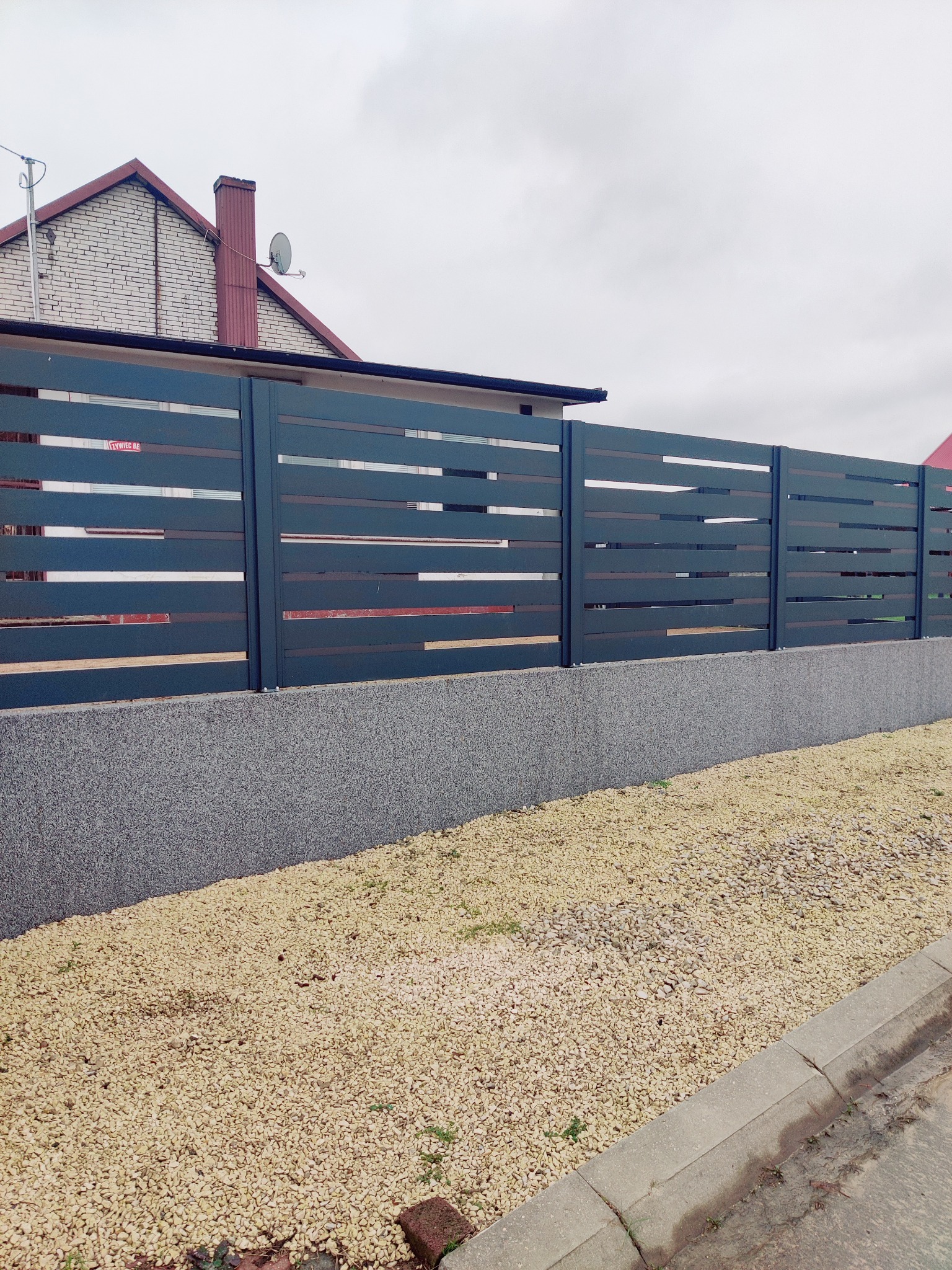 Nowoczesne, poziome przęsła ogrodzeniowe w kolorze grafitowym, zamontowane na betonowej podmurówce. Tło stanowi budynek mieszkalny i wysypany kamień ozdobny.