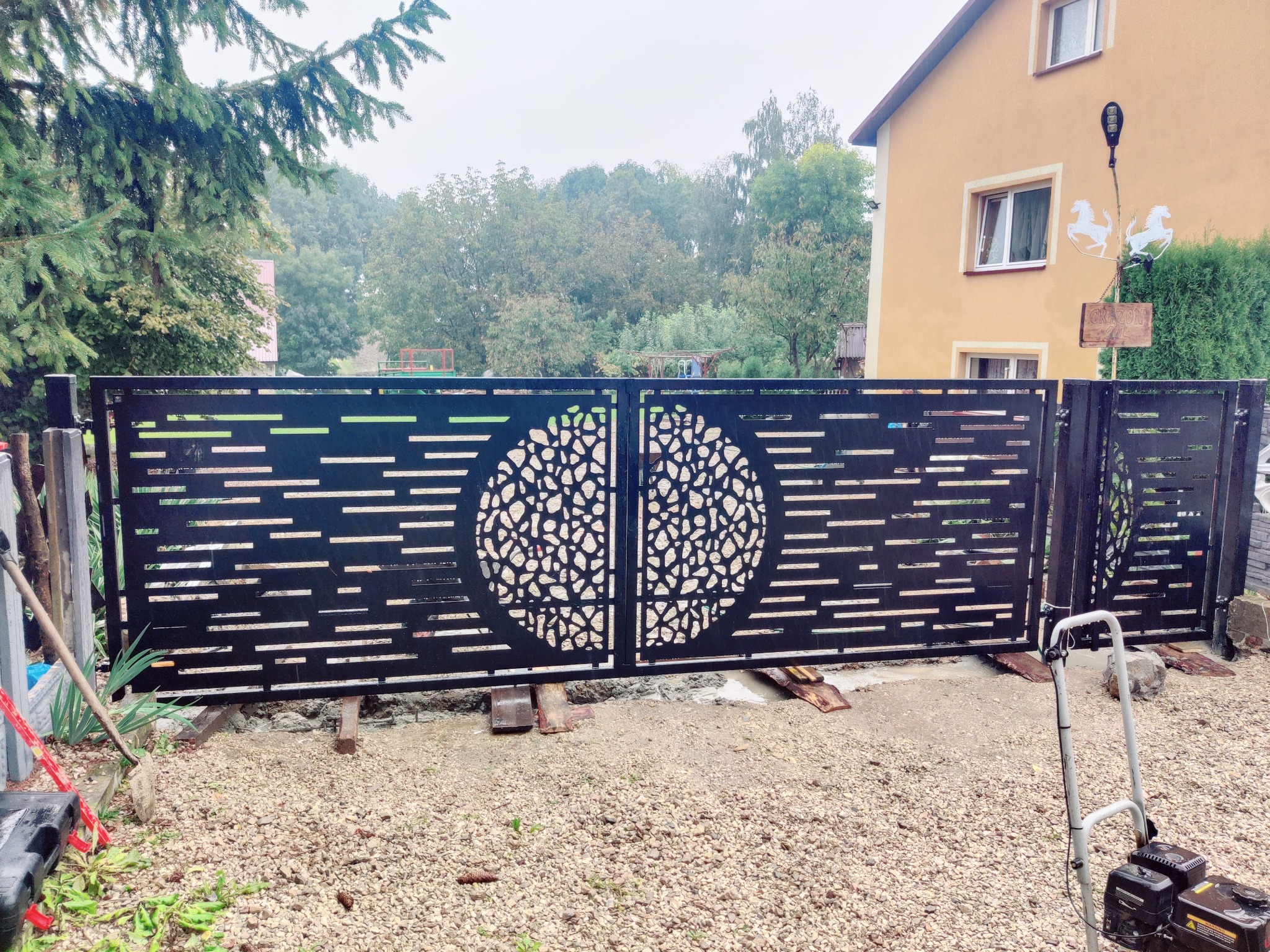 Nowoczesna, czarna brama wjazdowa z ażurowym wzorem koła i poziomych linii, zamontowana na posesji z widocznym domem w tle. Na pierwszym planie narzędzia.