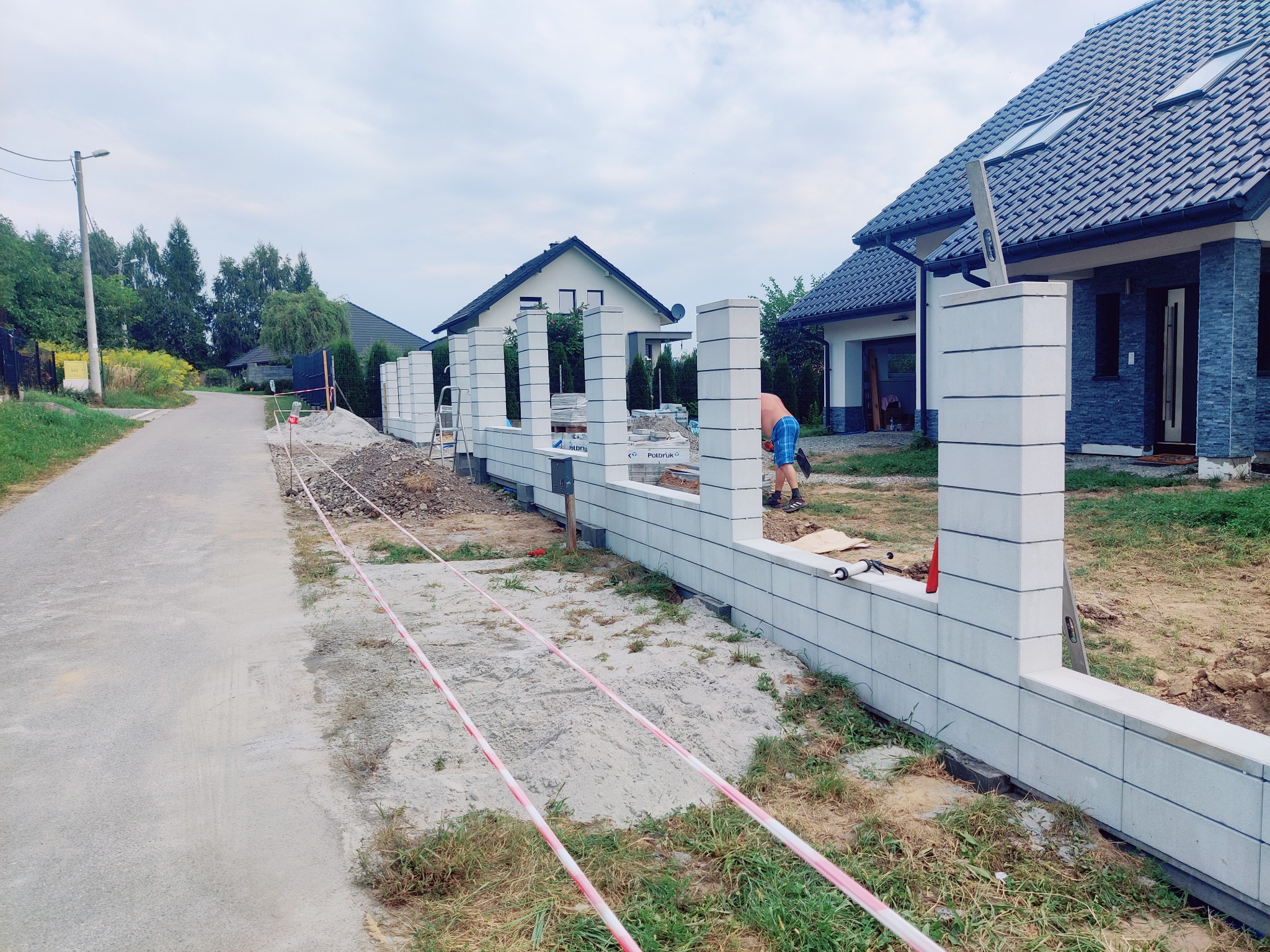 Wznoszenie ogrodzenia z prefabrykatów betonowych na posesji. Widoczne słupki i fragmenty muru, teren zabezpieczony taśmą. W tle domy jednorodzinne.