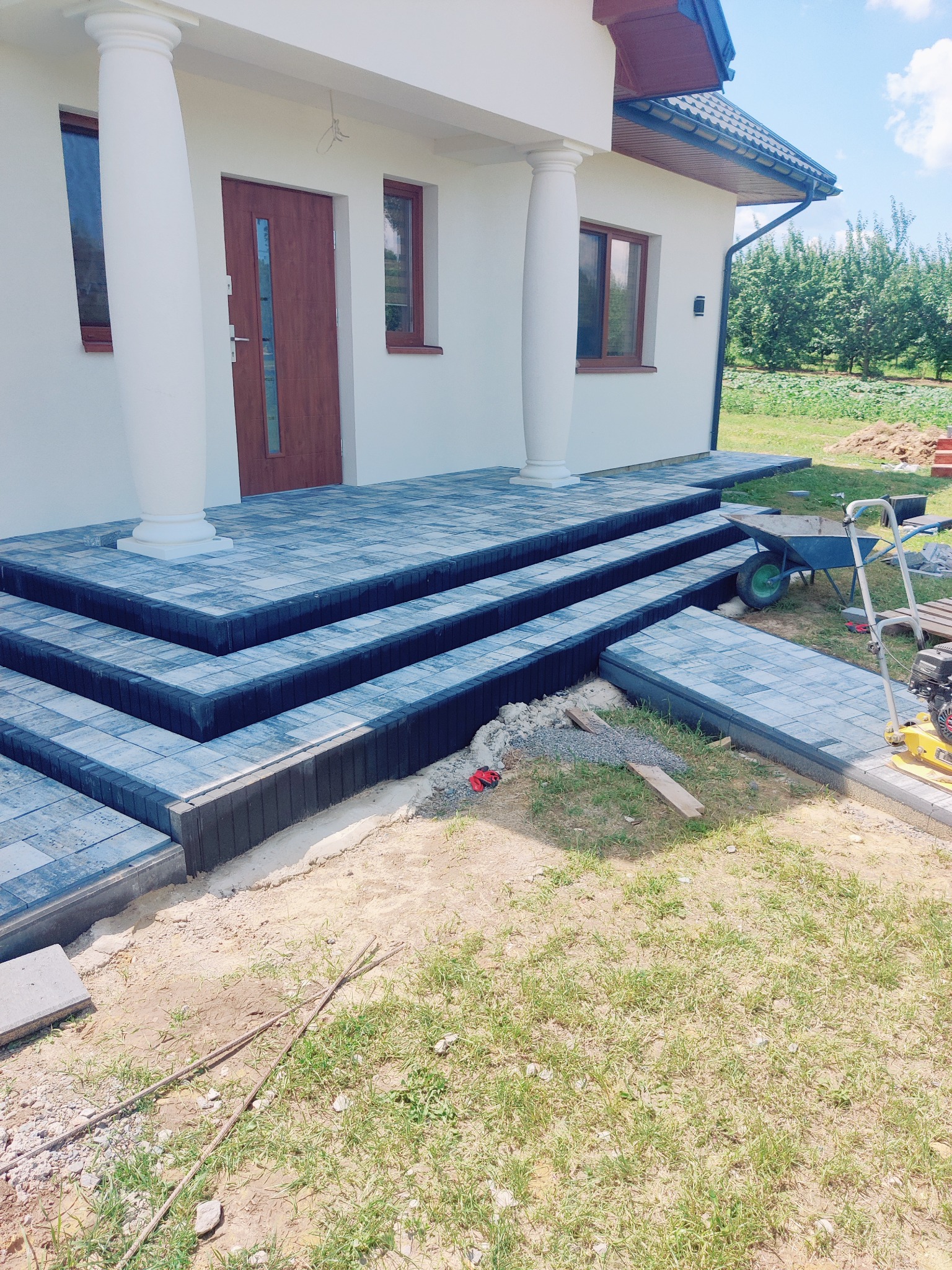 Ułożone schody wejściowe z kostki brukowej w odcieniach szarości, prowadzące do domu z kolumnami. Widoczne narzędzia brukarskie i elementy ogrodu.