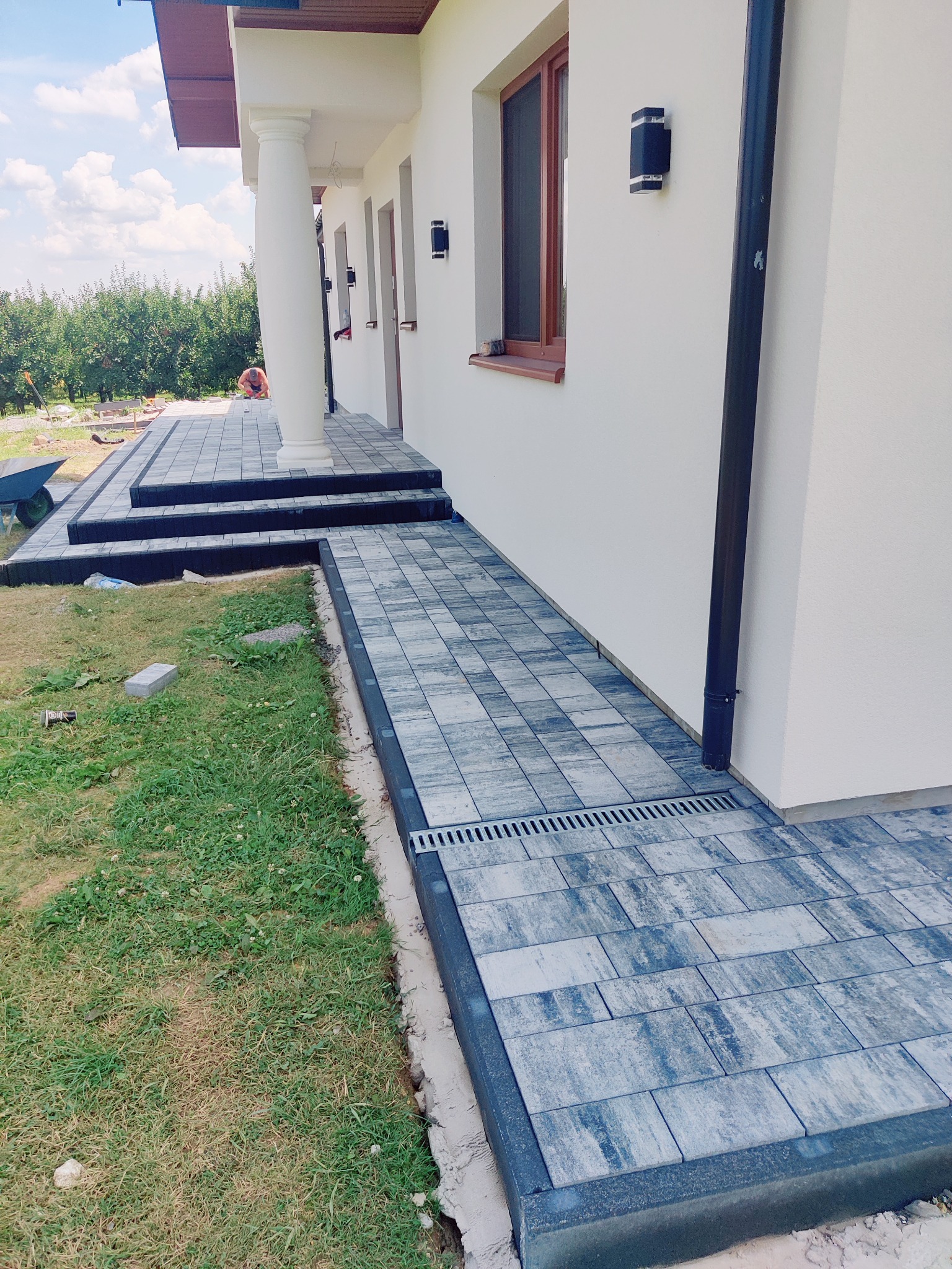 Ułożona kostka brukowa wzdłuż domu z kolumnami, widoczny system odprowadzania wody. Prace brukarskie na etapie wykończenia, widoczne narzędzia i materiały.