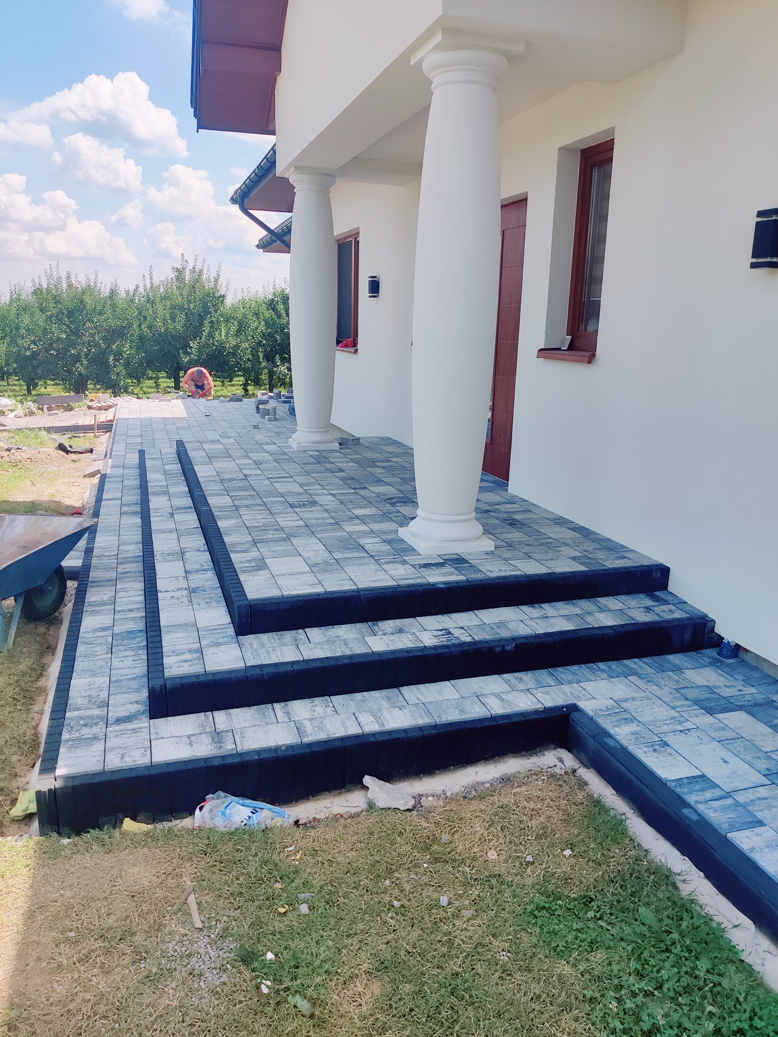 Ułożona kostka brukowa na schodach i chodniku przed domem z kolumnami. Widoczne szare i czarne kostki, trawnik i drzewa w tle. Częściowo widoczny wózek budowlany.