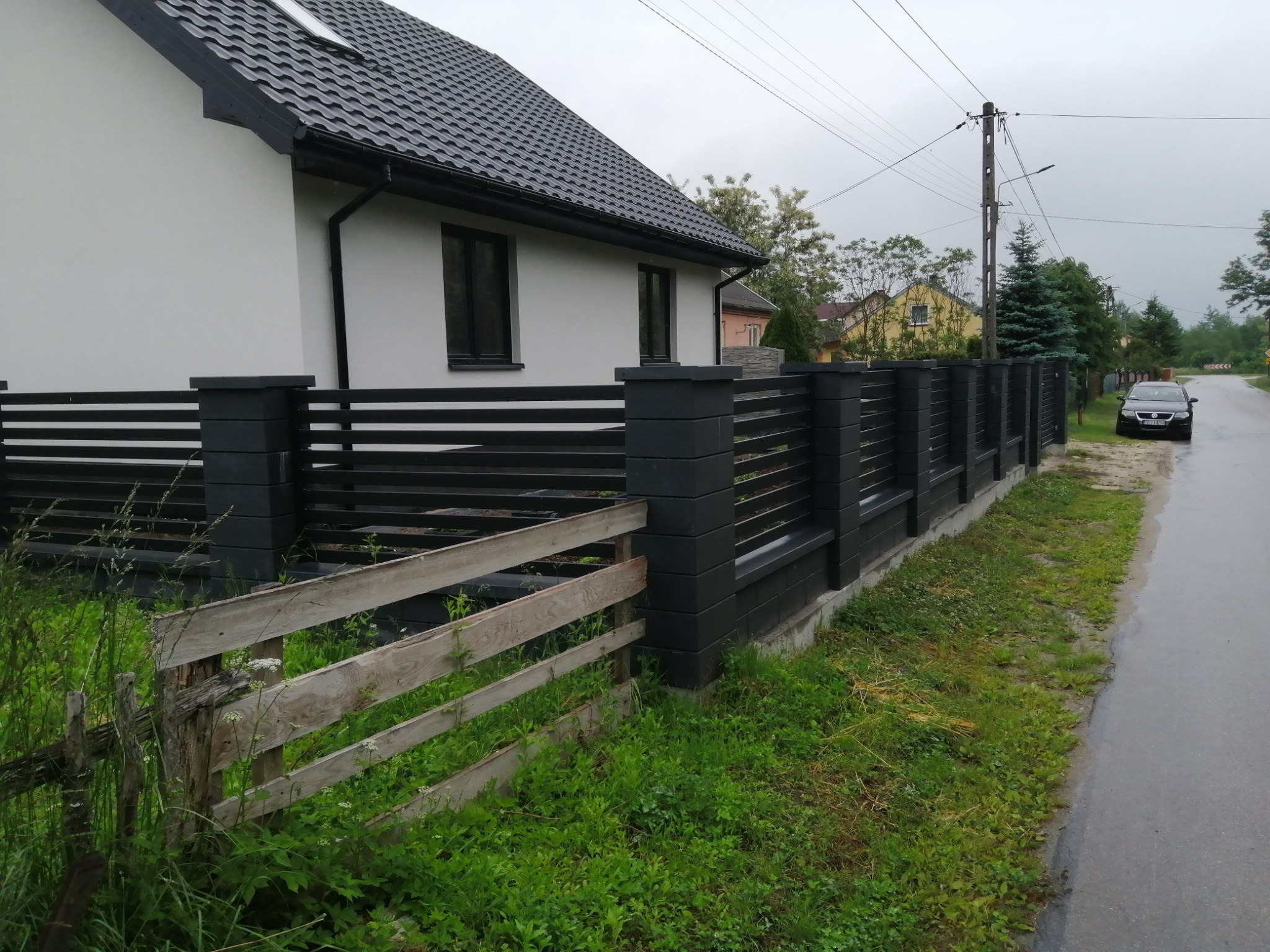 Nowoczesne ogrodzenie panelowe w kolorze grafitowym przylegające do białego domu z ciemną dachówką, z fragmentem starego, drewnianego płotu na pierwszym planie i zaparkowanym samochodem w tle.