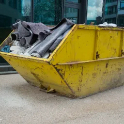 Żółty kontener na śmieci budowlane, częściowo wypełniony gruzem i przykryty szarą filcową tkaniną, stoi na chodniku na tle nowoczesnego budynku biurowego z szklaną fasadą.