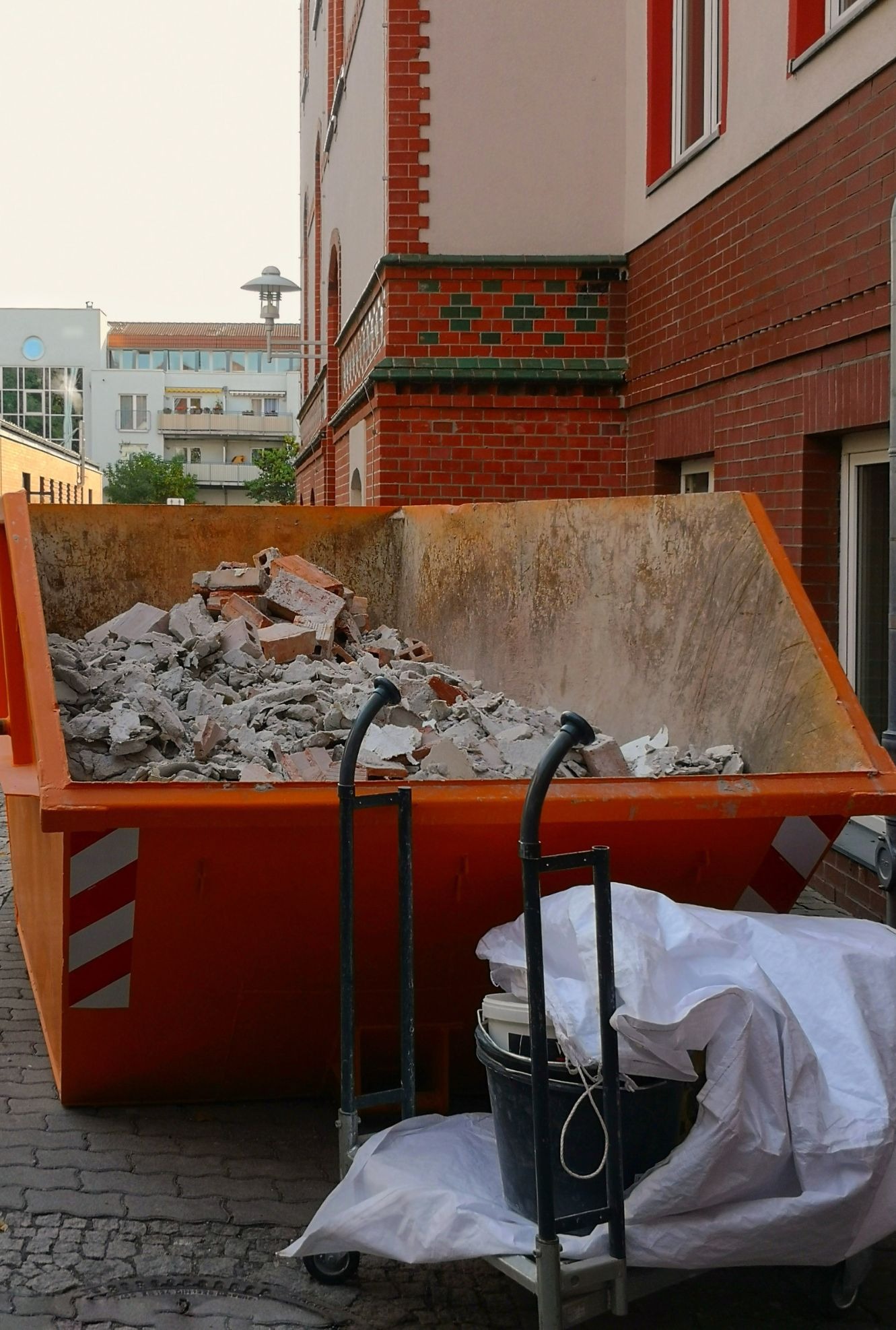 Pomarańczowy kontener wypełniony gruzem budowlanym przed budynkiem z czerwonej cegły, obok wózek z wiadrem i przykryty plandeką.