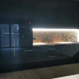 Czarna, minimalistyczna kuchnia z połyskującymi frontami, złotymi uchwytami i heksagonalną mozaiką na ścianie nad blatem.