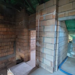 INSTAL-HOF Instalacje Elektryczne - Wnętrze budynku w trakcie budowy z widocznymi instalacjami kabli elektrycznych prowadzonymi po ścianach z cegły ceramicznej.