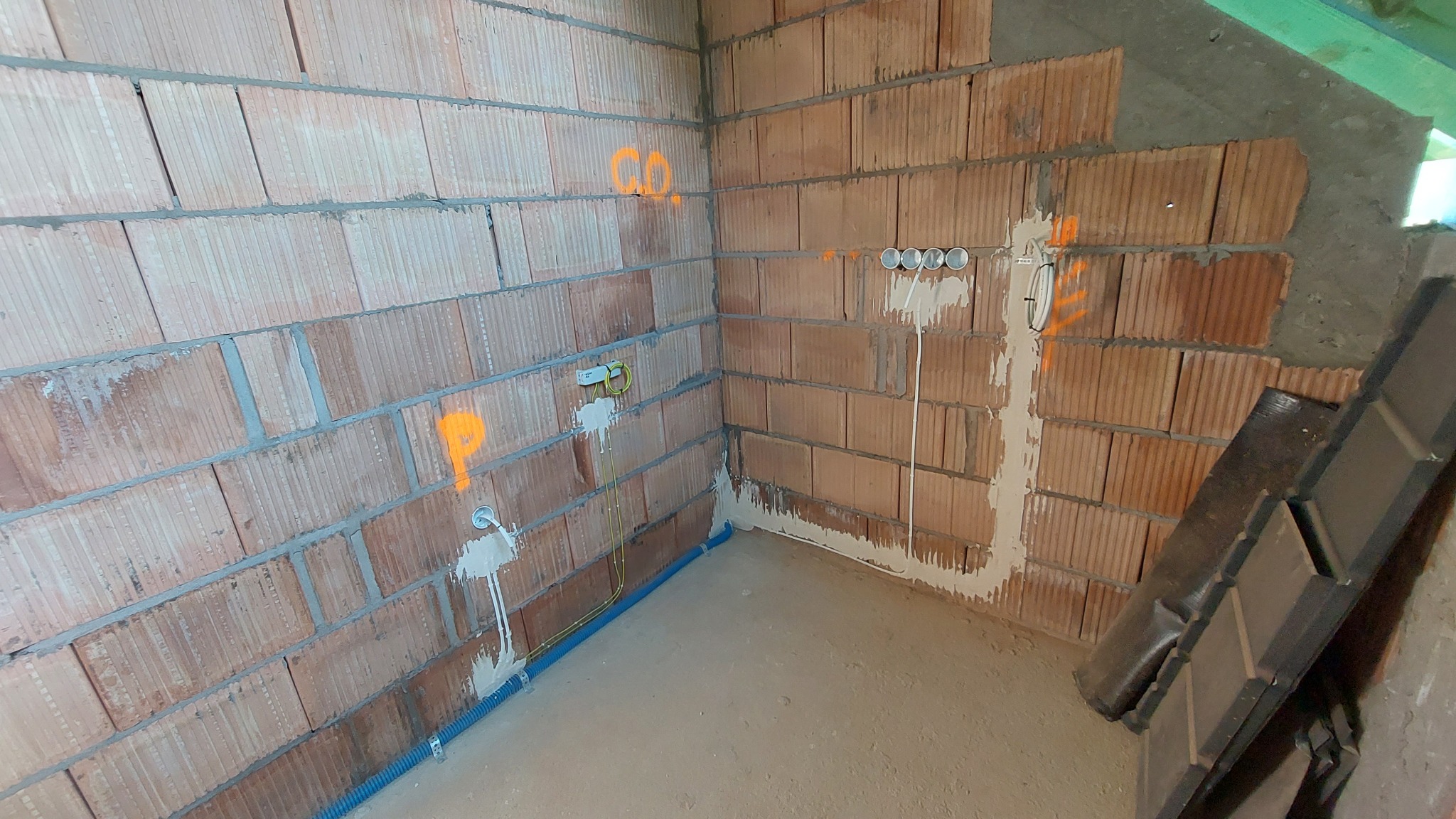 Wnętrze w trakcie prac: murowane ściany z widocznymi bruzdami pod kable, oznaczenia natryskowe w kolorze pomarańczowym, puszki instalacyjne i rury osłonowe w podłodze.