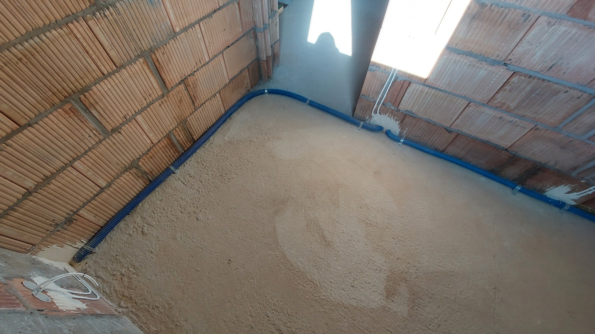 Niebieski karbowany przewód ochronny ułożony wzdłuż ściany z cegły dziurawki na surowym, szarym tynku w nowo budowanym pomieszczeniu, widoczne uchwyty mocujące.