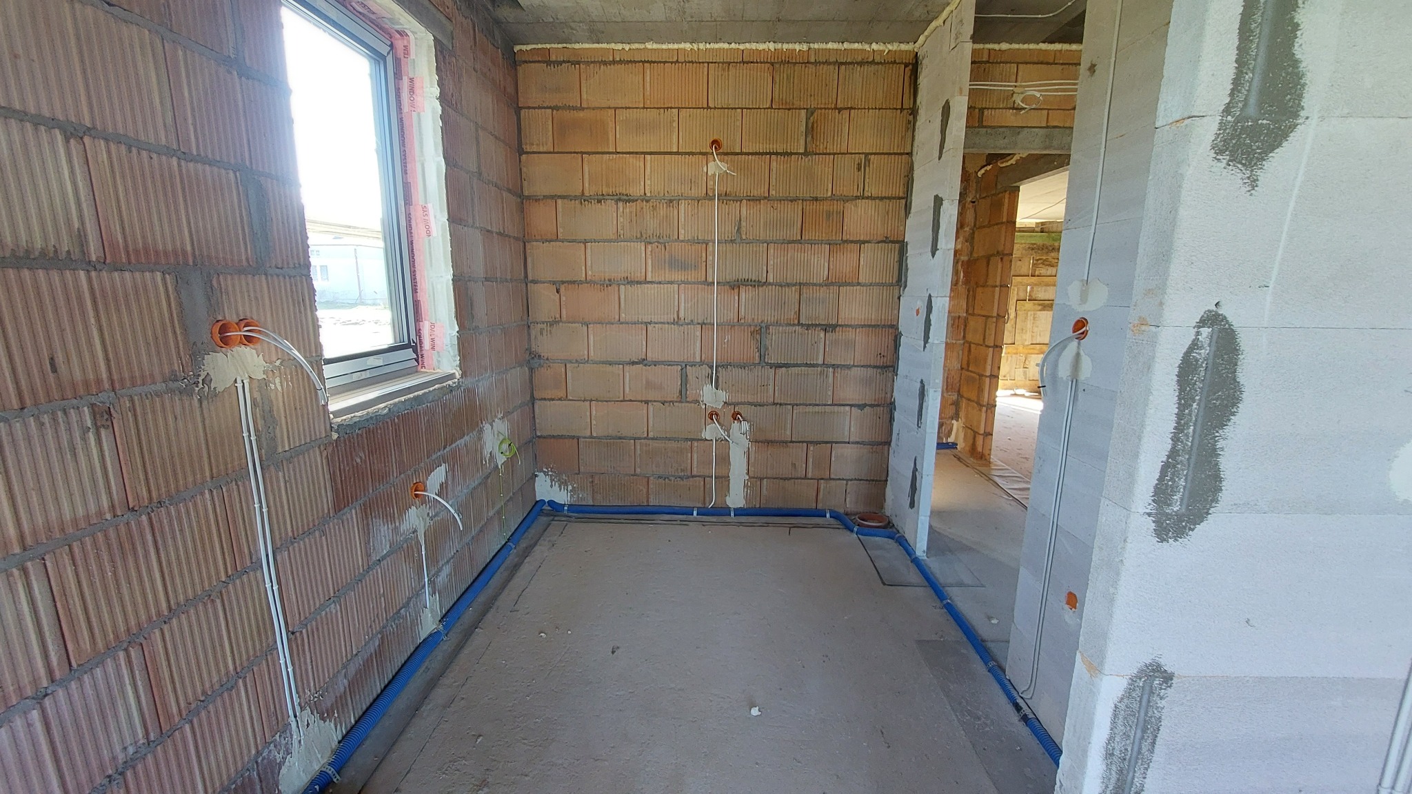 Wnętrze w stanie surowym z widocznymi przewodami elektrycznymi w ścianach z cegły i rurami ochronnymi na podłodze, przygotowane do dalszych prac wykończeniowych.