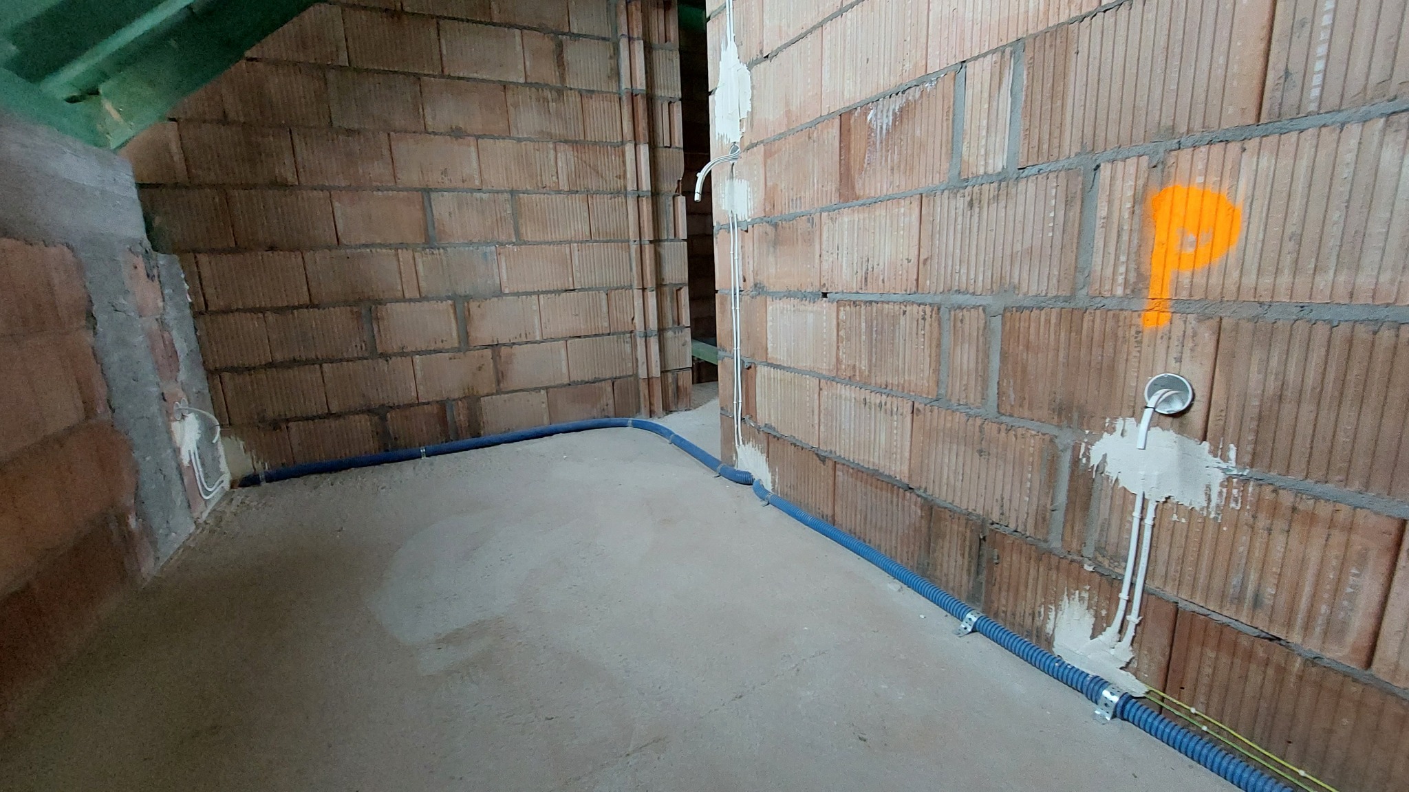 Wewnętrzne ściany z cegły z widoczną instalacją elektryczną: niebieska rura karbowana prowadzona wzdłuż podłogi, pionowe przewody w białej osłonie przymocowane do ściany, na ścianie pomarańczowa...