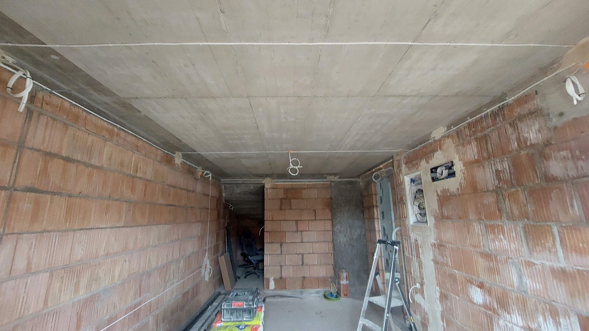 Surowy stan wnętrza z widoczną instalacją elektryczną: przewody poprowadzone pod sufitem, puszki elektryczne w ścianach z cegły, drabina rozstawiona przy ścianie.