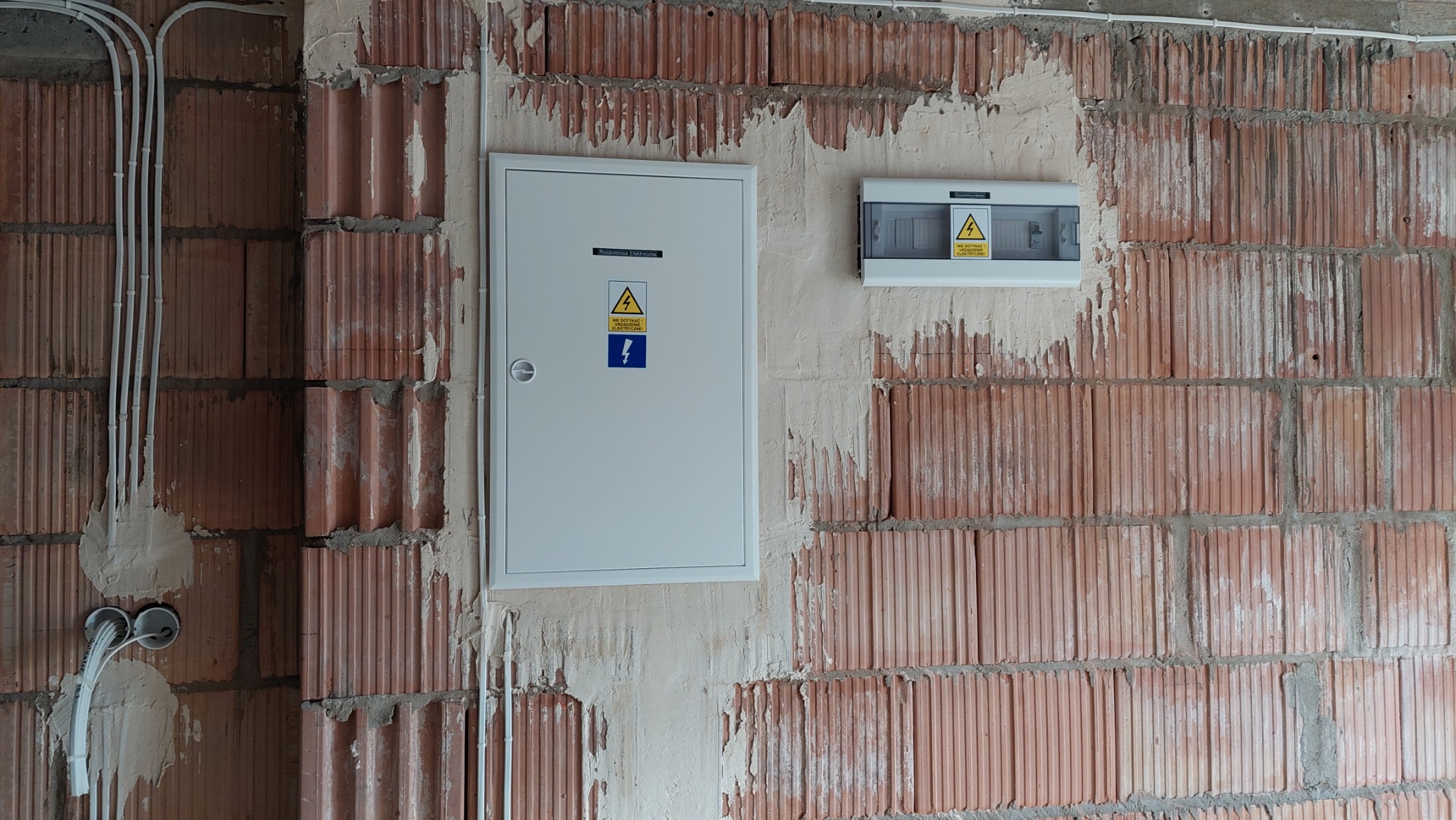 Dwie skrzynki elektryczne, jedna biała, druga szara, zamontowane na surowej ścianie z czerwonej cegły z widocznymi przewodami w białych rurkach