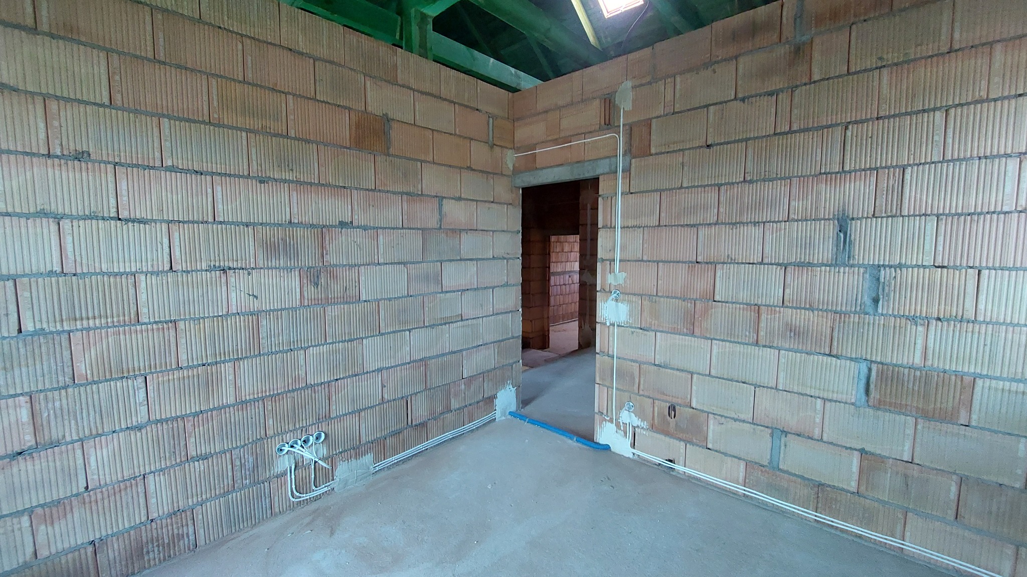Surowy mur z pustaków ceramicznych wewnątrz budynku, z widocznymi przewodami elektrycznymi w peszlach i puszkach, częściowo zamaskowanymi gipsem, na tle betonowej podłogi i zielonej konstrukcji dachu.