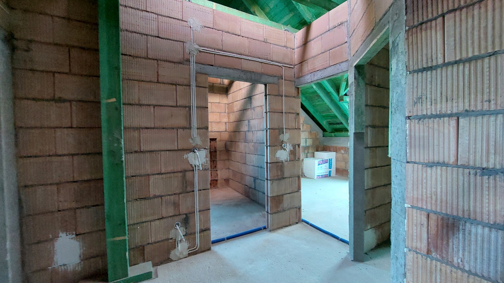 Surowy stan wnętrza z widoczną instalacją elektryczną w rurkach na ścianach z cegły dziurawki, nieotynkowane ściany, zielona więźba dachowa w tle, niebieskie rurki instalacyjne przy podłodze.
