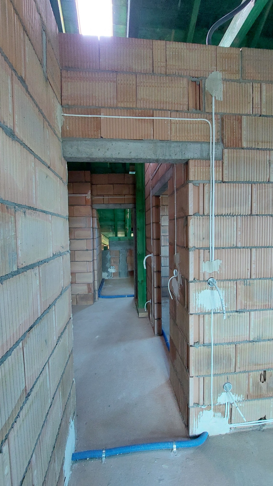 Wnętrze budynku w trakcie budowy z widocznymi przewodami elektrycznymi i rurami w ścianach z cegieł ceramicznych, surowy beton na podłodze, widoczny strop z belek drewnianych.