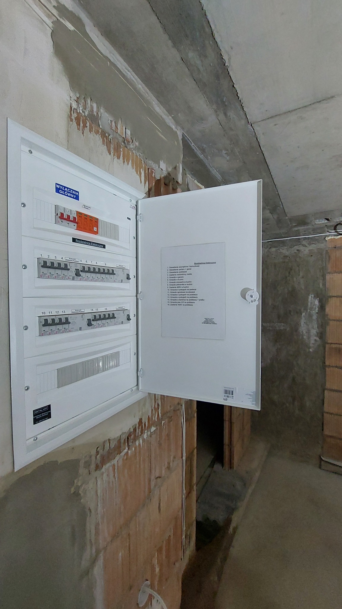 Otwarta skrzynka rozdzielcza z wyłącznikami i opisem, zamontowana na surowej ścianie z cegły w trakcie remontu.