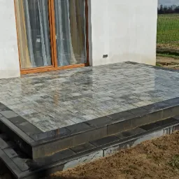 Nowo ułożony taras z kostki brukowej w odcieniach szarości i czerni, z dwoma stopniami, przylegający do białej ściany budynku z drewnianymi drzwiami balkonowymi i widoczną anteną satelitarną w tle.